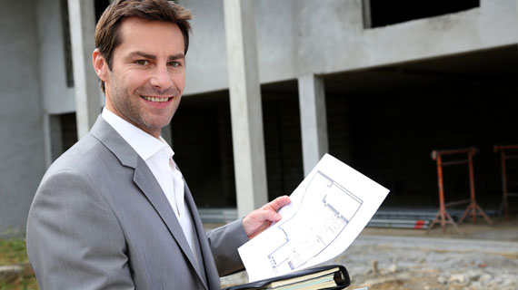 Ein lächelnder Mann im grauen Anzug steht vor einem Rohbau und hält Bauzeichnungen sowie einen Aktenordner in der Hand. Die offene Baustelle im Hintergrund zeigt unverputzte Betonpfeiler und Erdreich.