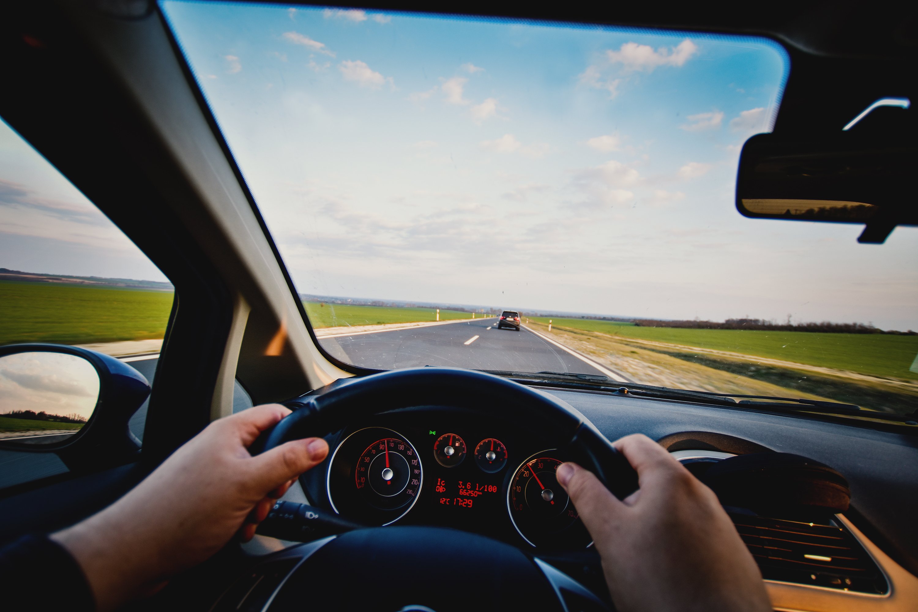 Blick aus der Fahrerperspektive auf eine Landstraße. Beide Hände liegen am Lenkrad, während ein weiteres Auto vorausfährt.