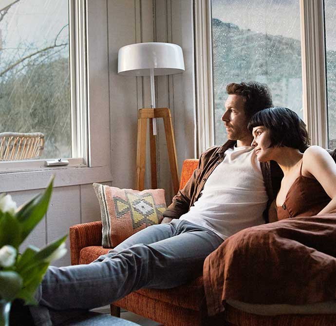 Ein Paar sitzt entspannt auf einem Sofa vor einem großen Fenster, während draußen Regen fällt. Die beiden wirken ruhig und geborgen.