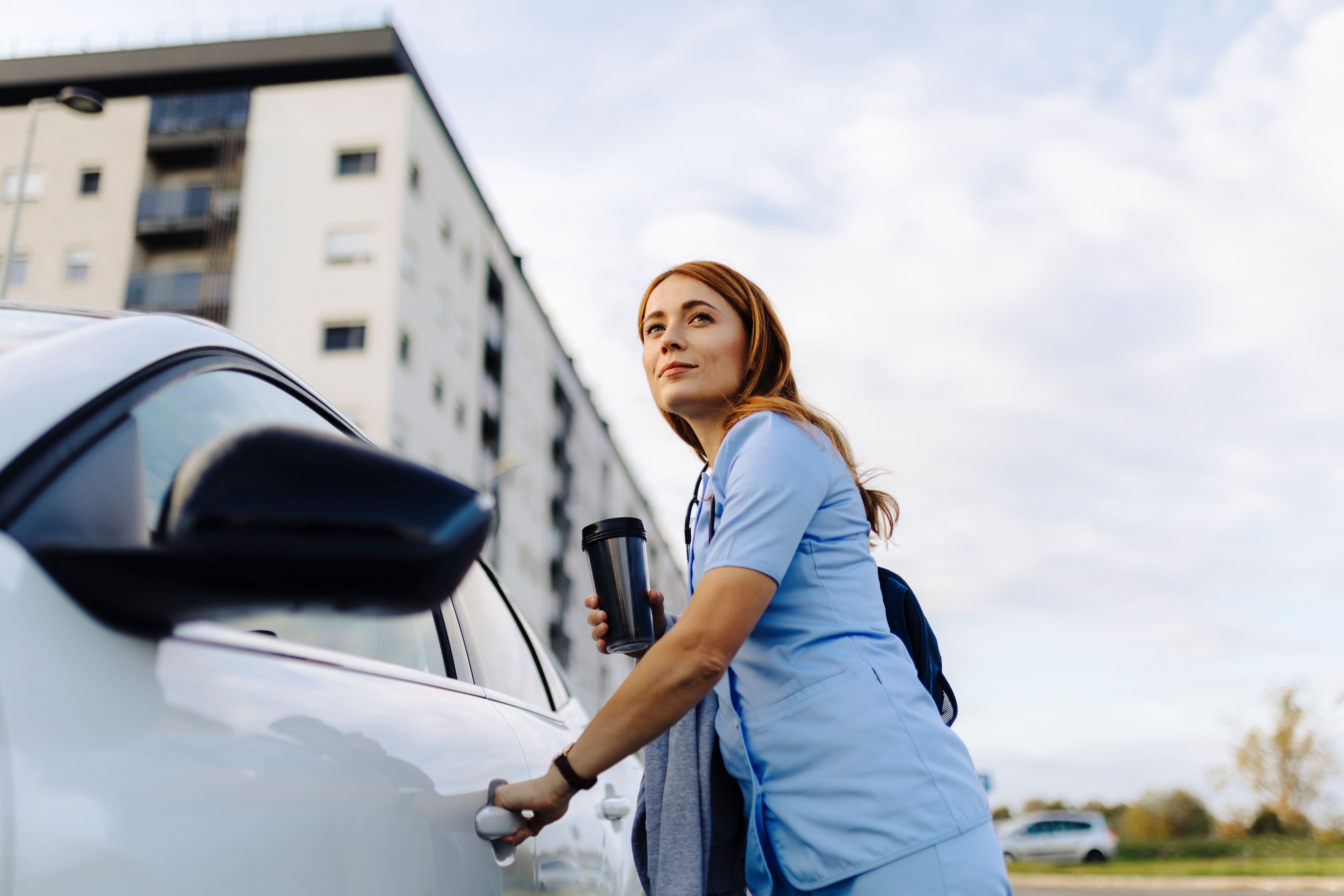 Eine Pflegekraft in blauer Arbeitskleidung öffnet die Tür eines weißen Autos und hält einen Kaffeebecher in der Hand.