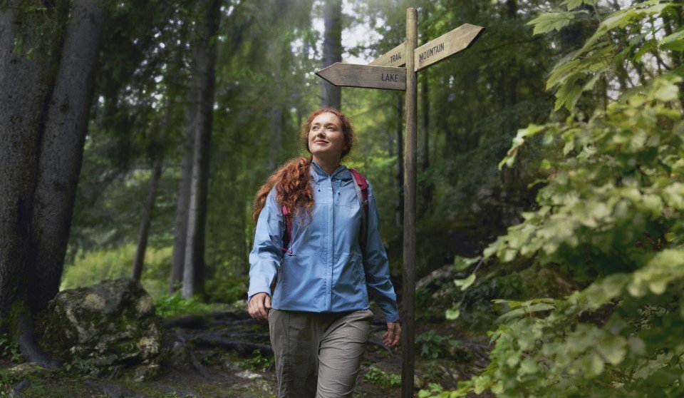 Junge Frau mit roten, lockigen Haaren und rotem Rucksack wandert in grauer Hose und blauer Outdoorjacke auf einem Waldweg an einem hölzernen Wegweiser vorbei, umgeben von dichtem, grünem Wald und moosbewachsenen Steinen.