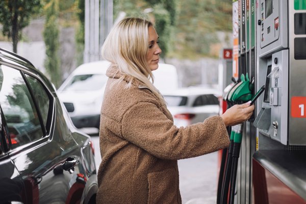 Eine Frau im braunen Mantel steht an einer Zapfsäule und hält eine grüne Zapfpistole, um ihr Auto zu betanken.