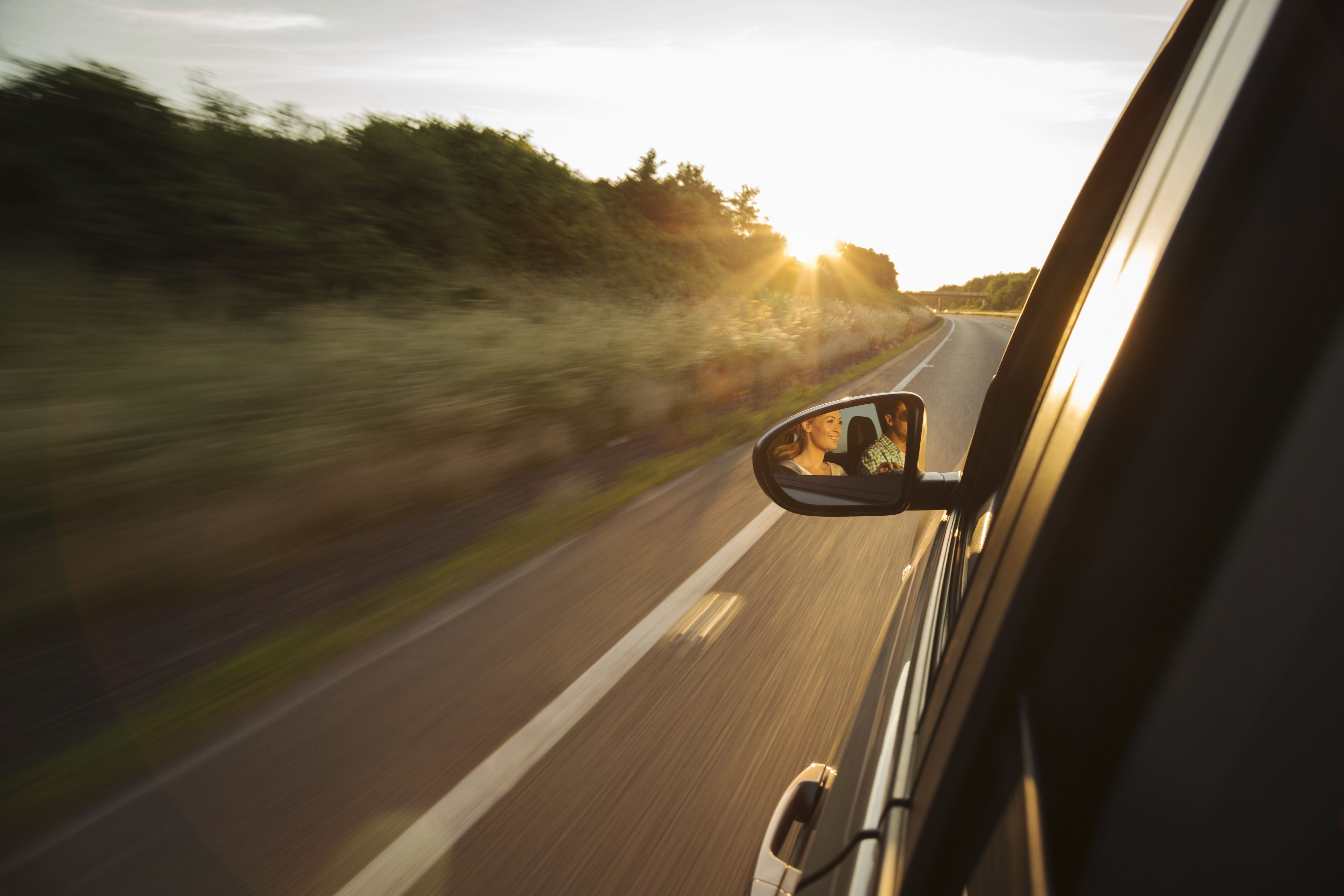 Ein Auto fährt bei tief stehender Sonne auf einer Landstraße. Im Außenspiegel sind zwei Personen im Fahrzeug zu sehen.