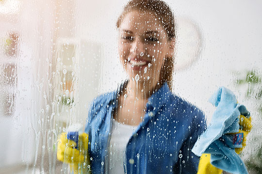 Eine lächelnde Frau mit gelben Gummihandschuhen reinigt eine Glasfläche. Wassertropfen und Reinigungsmittel sind auf dem Glas sichtbar, während sie ein Putztuch und eine Sprühflasche hält.