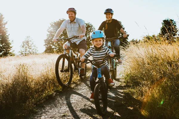 Familie mit Kind auf Fahrrädern fährt einen sonnigen Feldweg entlang, das Kind trägt einen blauen Helm und lacht.