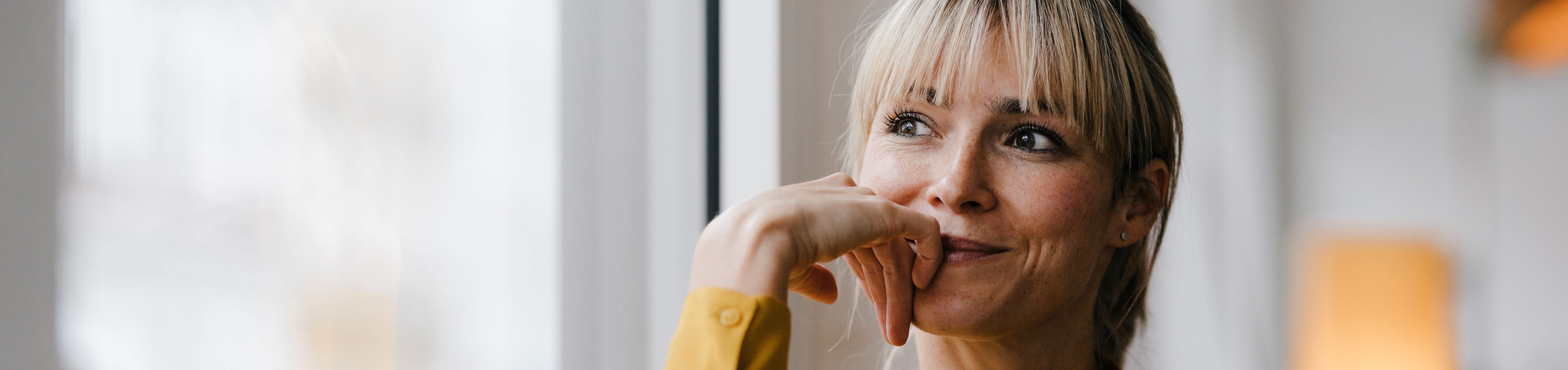 Zahnzusatzversicherung ohne Gesundheitsfragen Eine blonde Frau mit Pony und gelber Bluse sitzt an einem Fenster. Sie stützt ihr Kinn auf ihre Hand, lächelt leicht und blickt nachdenklich zur Seite.