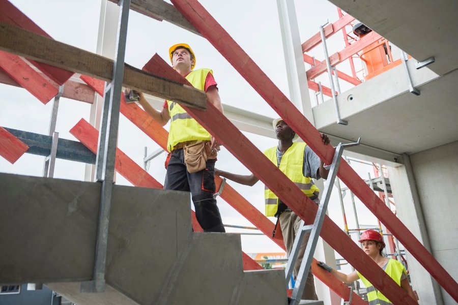 Drei Arbeiter mit Schutzwesten steigen eine Betontreppe auf einer Baustelle hinauf.