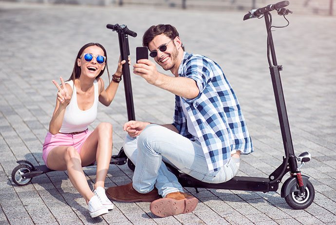 Ein junger Mann und eine Frau sitzen lachend auf dem Boden neben ihren E-Scootern und machen ein Selfie. Beide tragen Sonnenbrillen und wirken fröhlich.