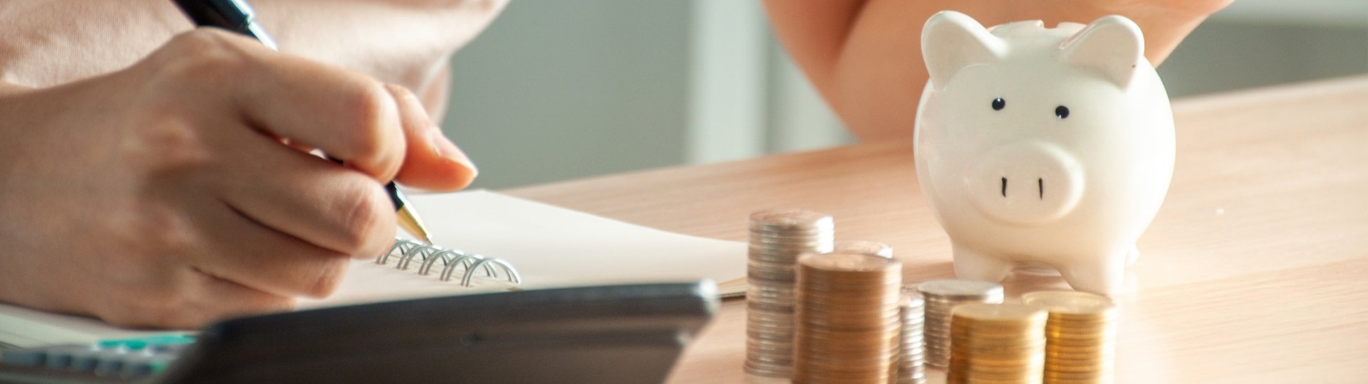 Eine Person macht sich Notizen zur Finanzplanung. Auf dem Tisch stehen ein Sparschwein und mehrere Stapel mit M&uuml;nzen als Symbol f&uuml;r Sparen und Altersvorsorge.