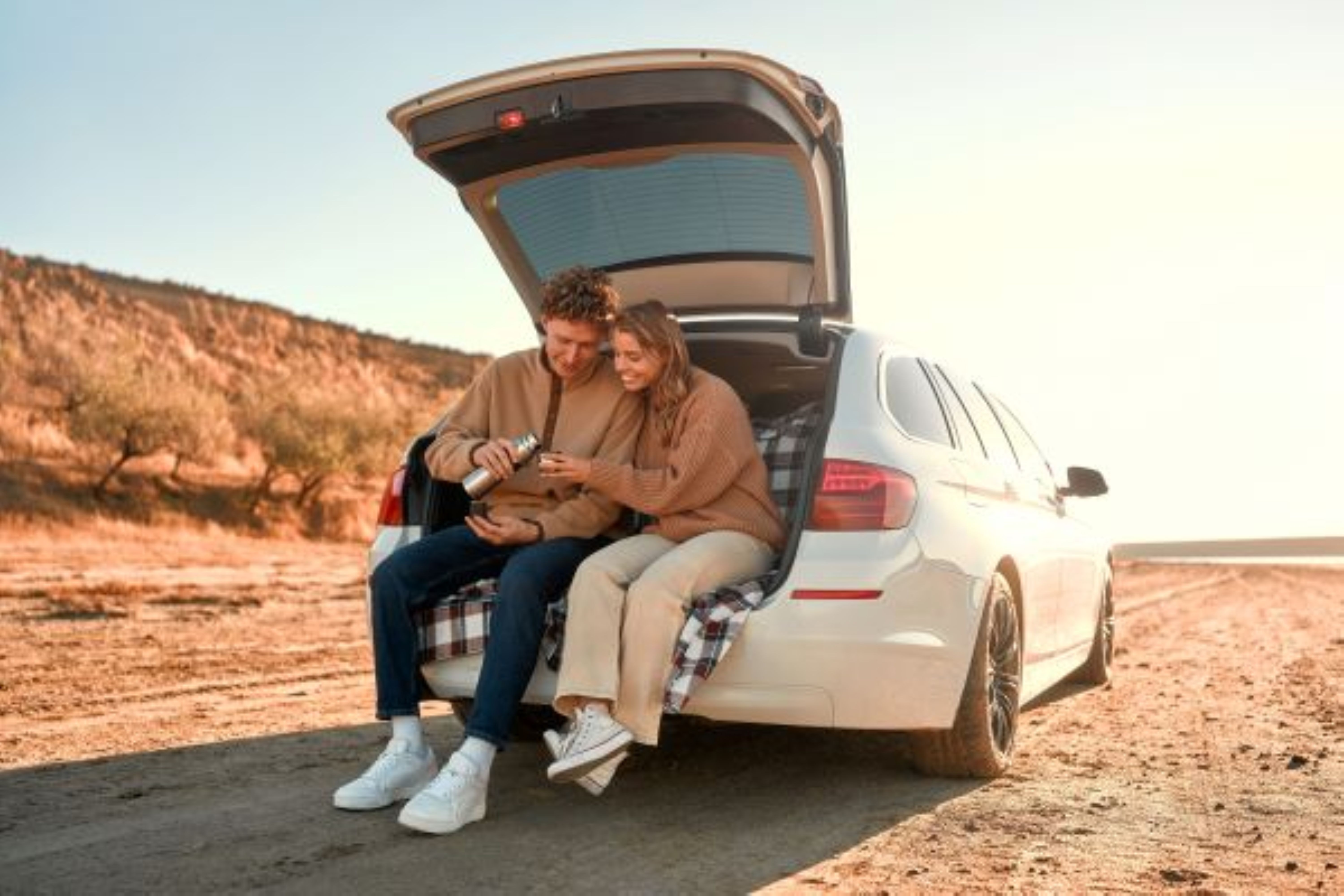 Ein junges Paar sitzt am geöffneten Kofferraum eines weißen Autos und macht eine Pause in einer sonnigen Landschaft.