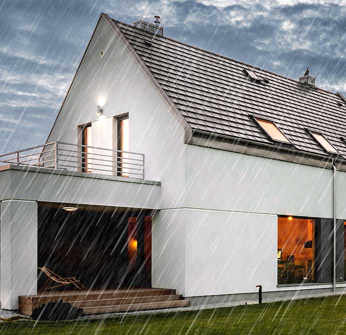 Modernes Einfamilienhaus mit Flachdach und großen Fenstern bei starkem Regen. Die Außenbeleuchtung ist eingeschaltet, das Haus wirkt stabil und gut geschützt.