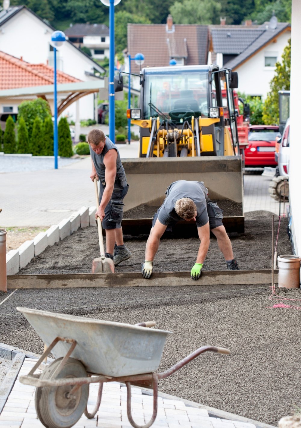 Zwei Bauarbeiter ebnen einen Schotterweg vor einem Haus, ein Radlader im Hintergrund.