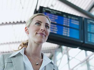 Eine blonde Geschäftsfrau in einem hellen Blazer steht an einem Flughafen. Im Hintergrund sind große Anzeigetafeln mit Abfluginformationen zu sehen.