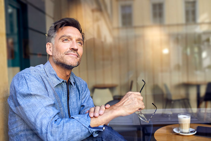 Mann sitzt im Café am Fenster und blickt nach draußen, hält eine Brille in der Hand