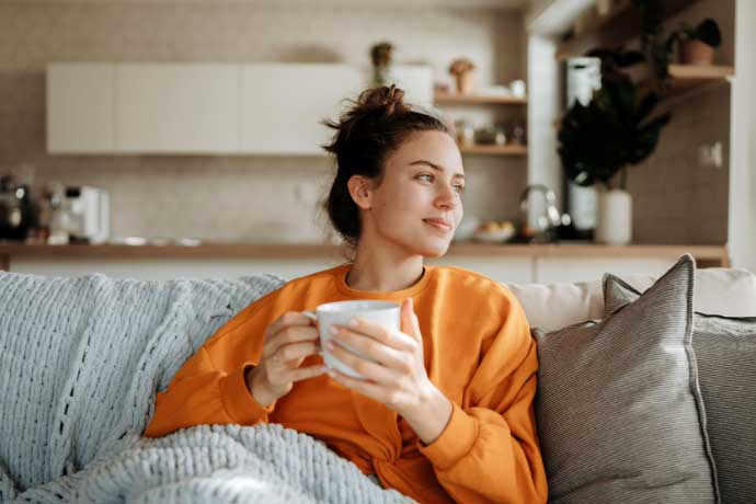 Junge Frau in orangefarbenem Pullover sitzt entspannt auf einem Sofa, hält eine Tasse in beiden Händen und blickt nachdenklich aus dem Fenster. Im Hintergrund ist eine moderne Küche zu sehen.