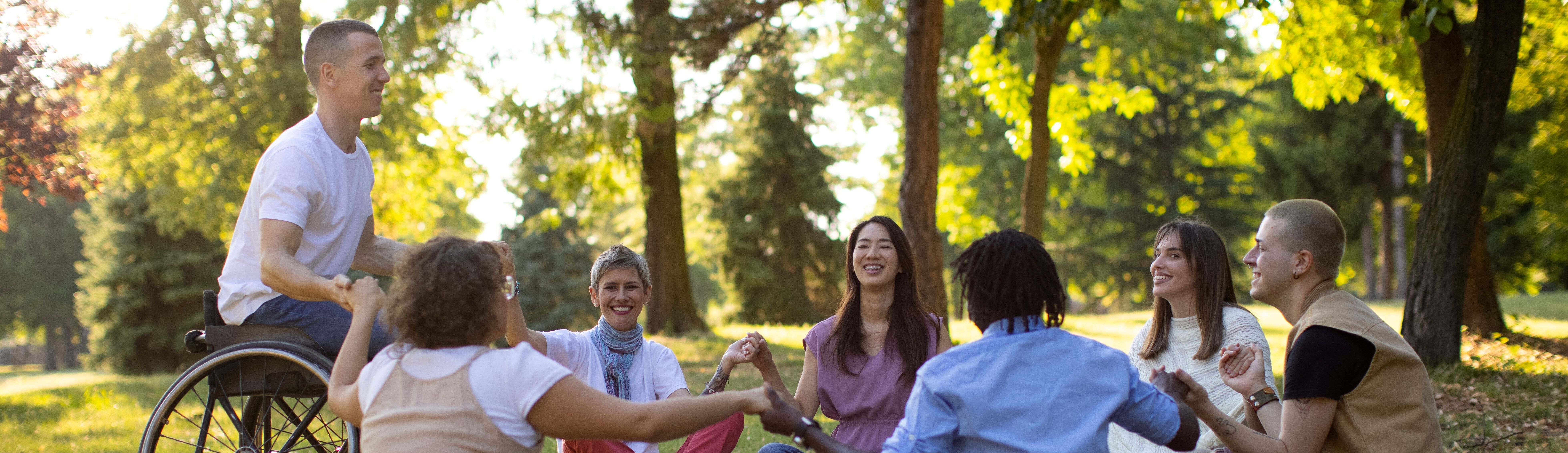 Mitarbeiter sitzen gemeinsam im Park und halten sich an den H&auml;nden. Eine Gruppe verschiedener Personen tauscht sich aus und wirkt dabei offen und verbunden.