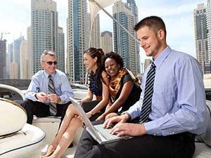  Eine Gruppe internationaler Geschäftsleute entspannt auf einer Jacht mit Blick auf eine moderne Skyline. Ein Mann im Vordergrund arbeitet an einem Laptop, während die anderen lachen und sich unterhalten.