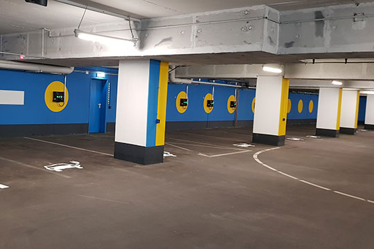 Tiefgaragenparkplätze mit mehreren Ladestationen für Elektroautos. Die Wände sind blau und gelb gestaltet, und die Parkflächen sind für E-Fahrzeuge reserviert.