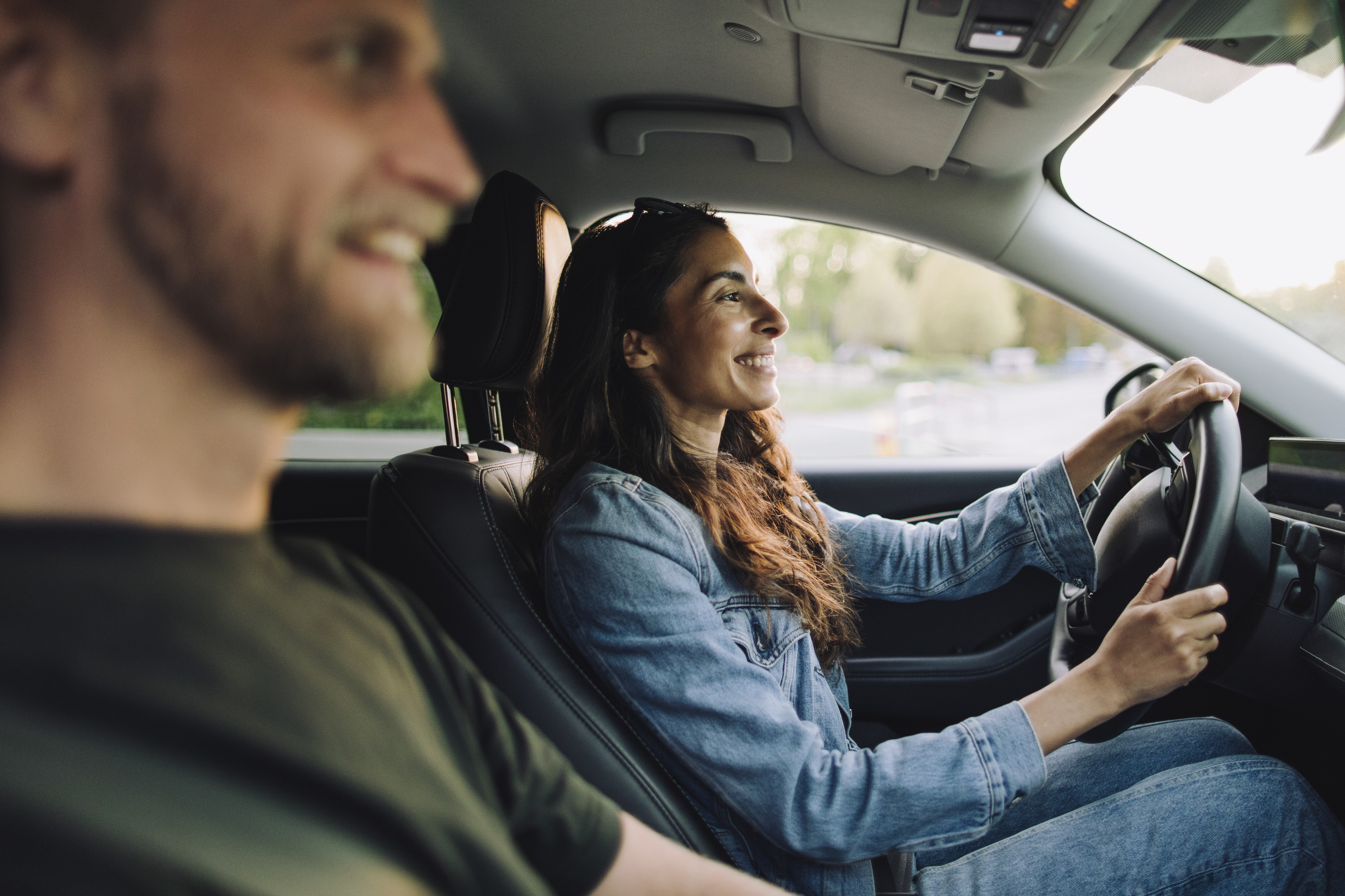 Eine lächelnde Frau fährt ein Auto, während ein Mann auf dem Beifahrersitz mitfährt.