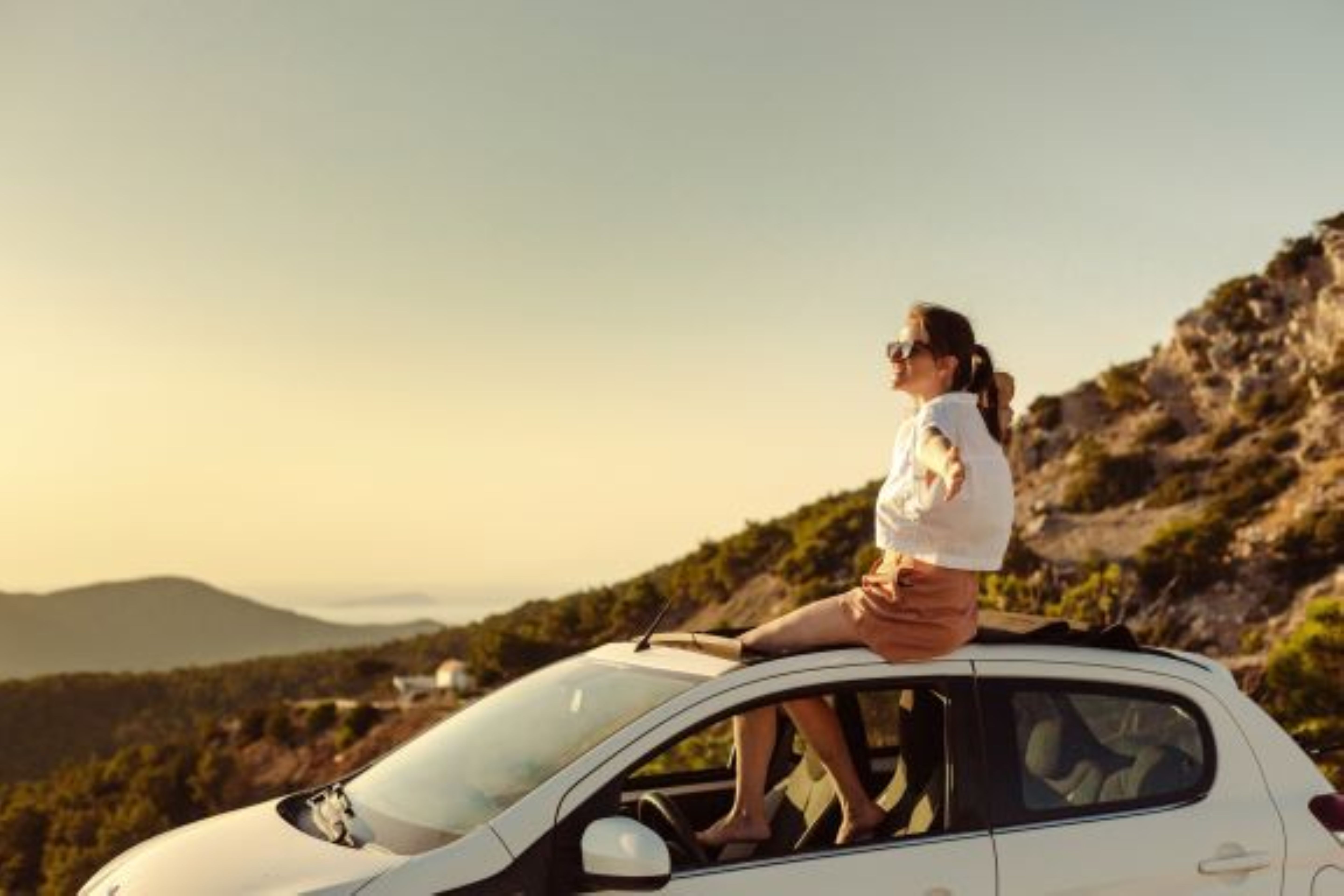Eine Frau mit Sonnenbrille sitzt auf dem Dach eines weißen Autos vor einer bergigen Landschaft im warmen Abendlicht.