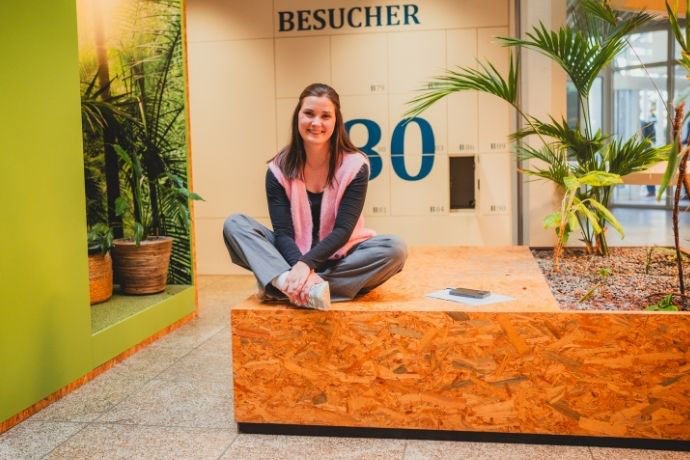 Trainee sitzt im Schneidersitz in grünem AXA Foyer und lacht