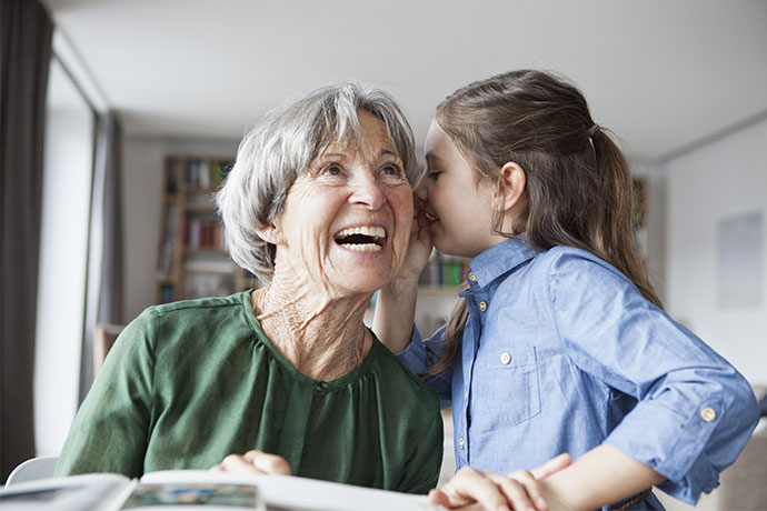Mädchen flüstert einer Seniorin etwas in das linke Ohr. Diese lacht.