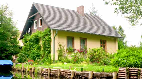 Ein Einfamilienhaus mit gelber Fassade und begrüntem Dachgiebel steht an einem Kanal. Der Garten ist dicht bepflanzt mit Blumen und Sträuchern, ein Holzbalken-Ufer schützt das Grundstück vor dem Wasserlauf.