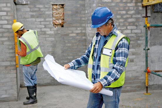 Ein Bauleiter mit blauer Schutzhelmkappe und Sicherheitsweste betrachtet aufmerksam Baupläne auf einer Baustelle mit unverputzten Ziegelwänden. Im Hintergrund misst ein weiterer Bauarbeiter mit Wasserwaage eine Wand.