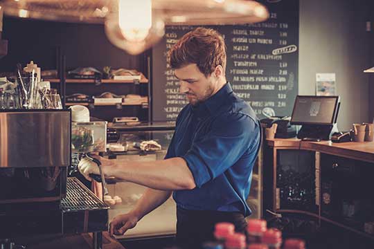 Ein Barista mit dunkelblauem Hemd bereitet an einer Siebträgermaschine Kaffee zu. Im Hintergrund sind eine Tafel mit Getränkepreisen, eine Kasse und Regale mit Backwaren zu sehen.